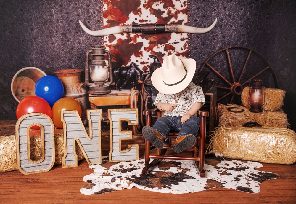 Toddler in cowboy hat sits in rocking chair surrounded by hay bales, ONE marquee letters, and western decor for first birthday.