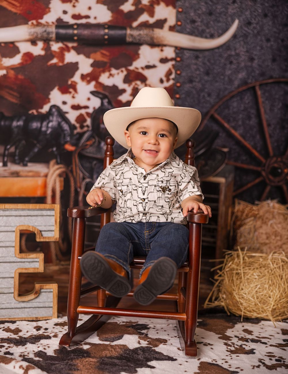Smiling baby boy in white cowboy hat and jeans sits in rocking chair with western ranch backdrop.