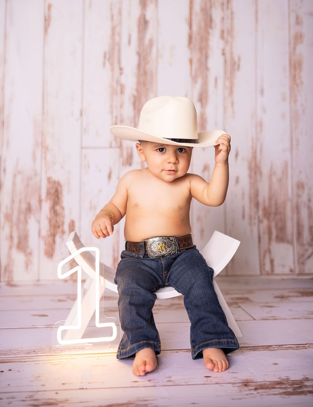Shirtless toddler wearing cowboy hat and jeans poses next to number one sign for first birthday photo.