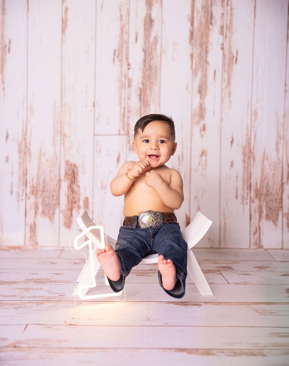 Smiling baby boy in jeans and belt sitting on a white rocking horse against a rustic white wood backdrop.