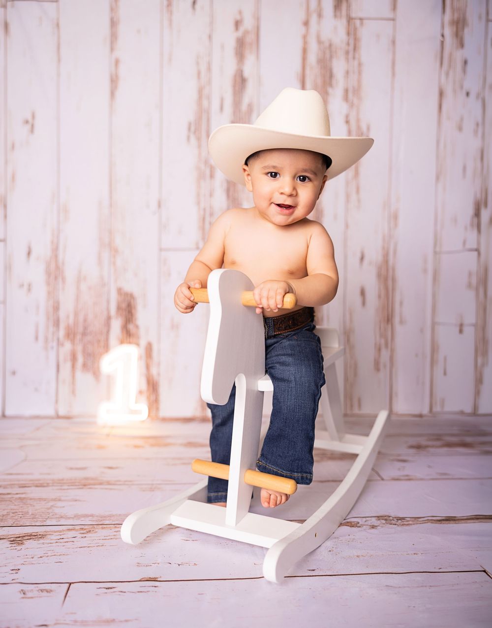 Smiling baby boy in cowboy hat and jeans standing on white wooden rocking horse toy.