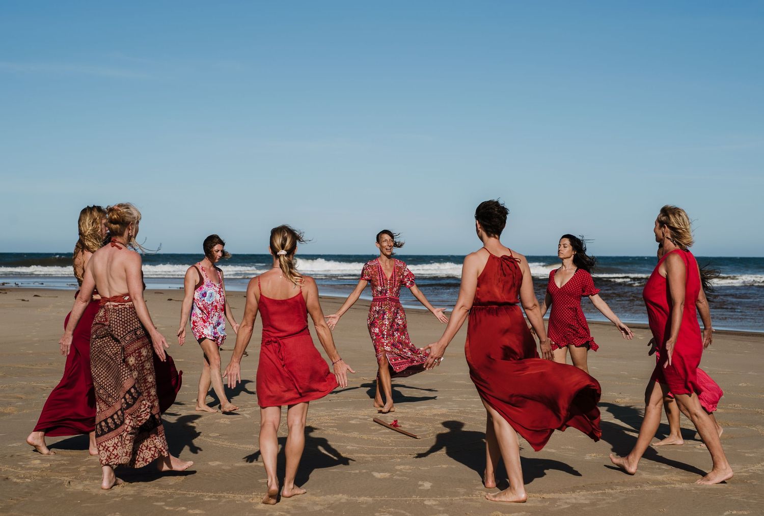 Dancers in flowing red dresses perform choreographed movements on a sandy beach.