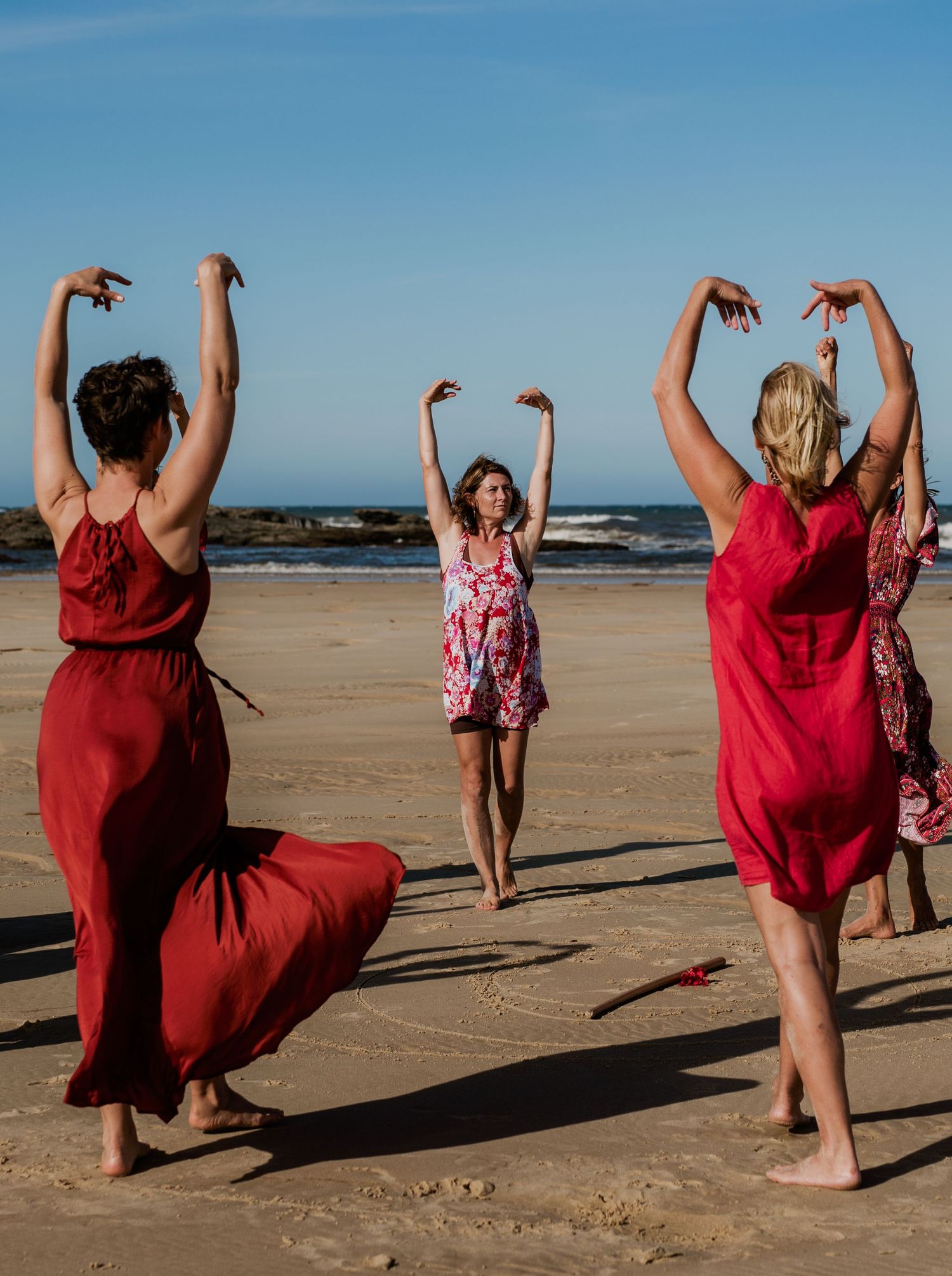 Group performs synchronized dance moves wearing red dresses on a sunny beach.