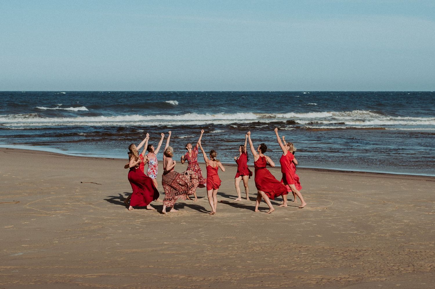 Dancers in red outfits pose together on a beach with ocean waves in the background.