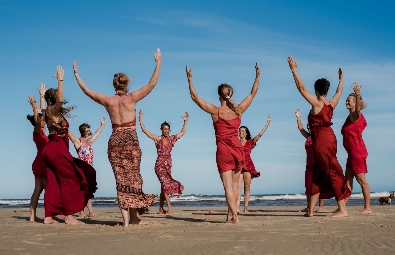 Dancers raise their arms in unison while performing on a sunny beach in red costumes.
