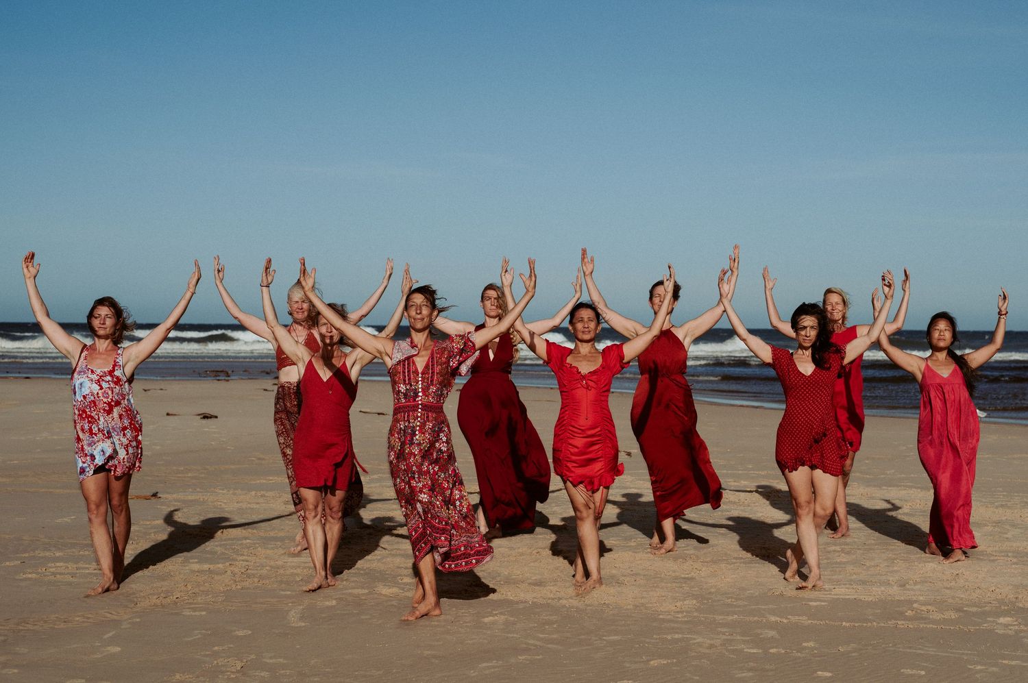 Group wearing red dresses jumping and posing together on sandy beach under blue sky.