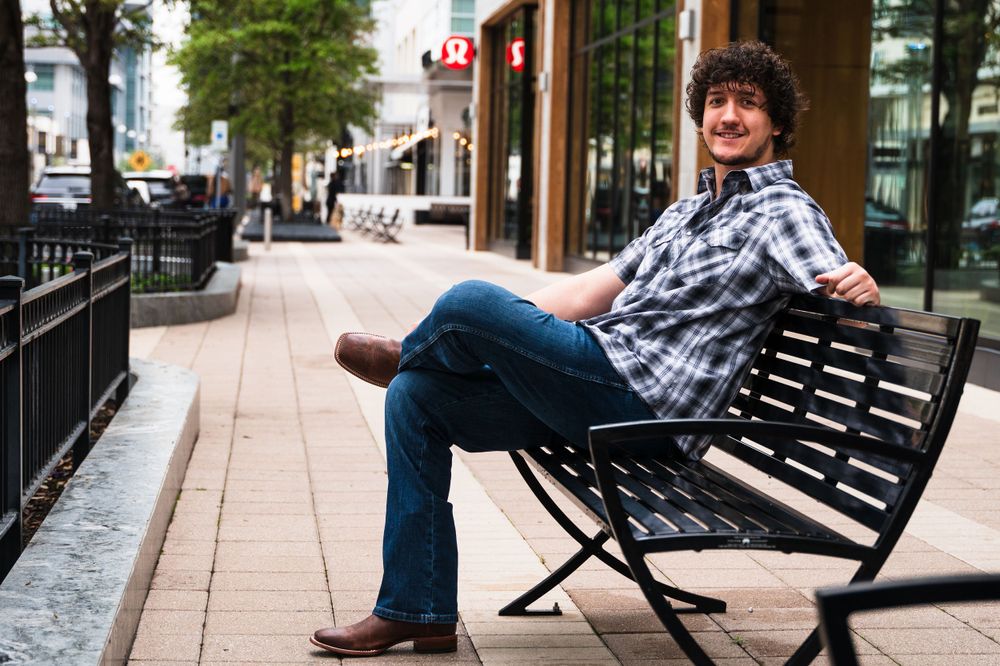 Casual portrait of person relaxing on bench in plaid shirt and jeans along urban sidewalk.