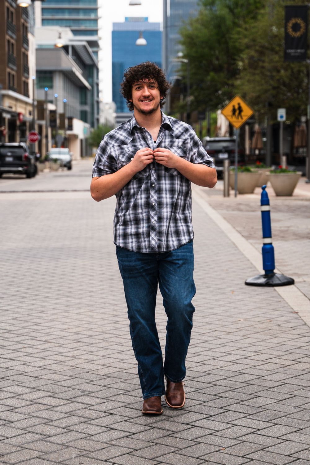 Person in plaid shirt walking down city street with tall buildings in background.
