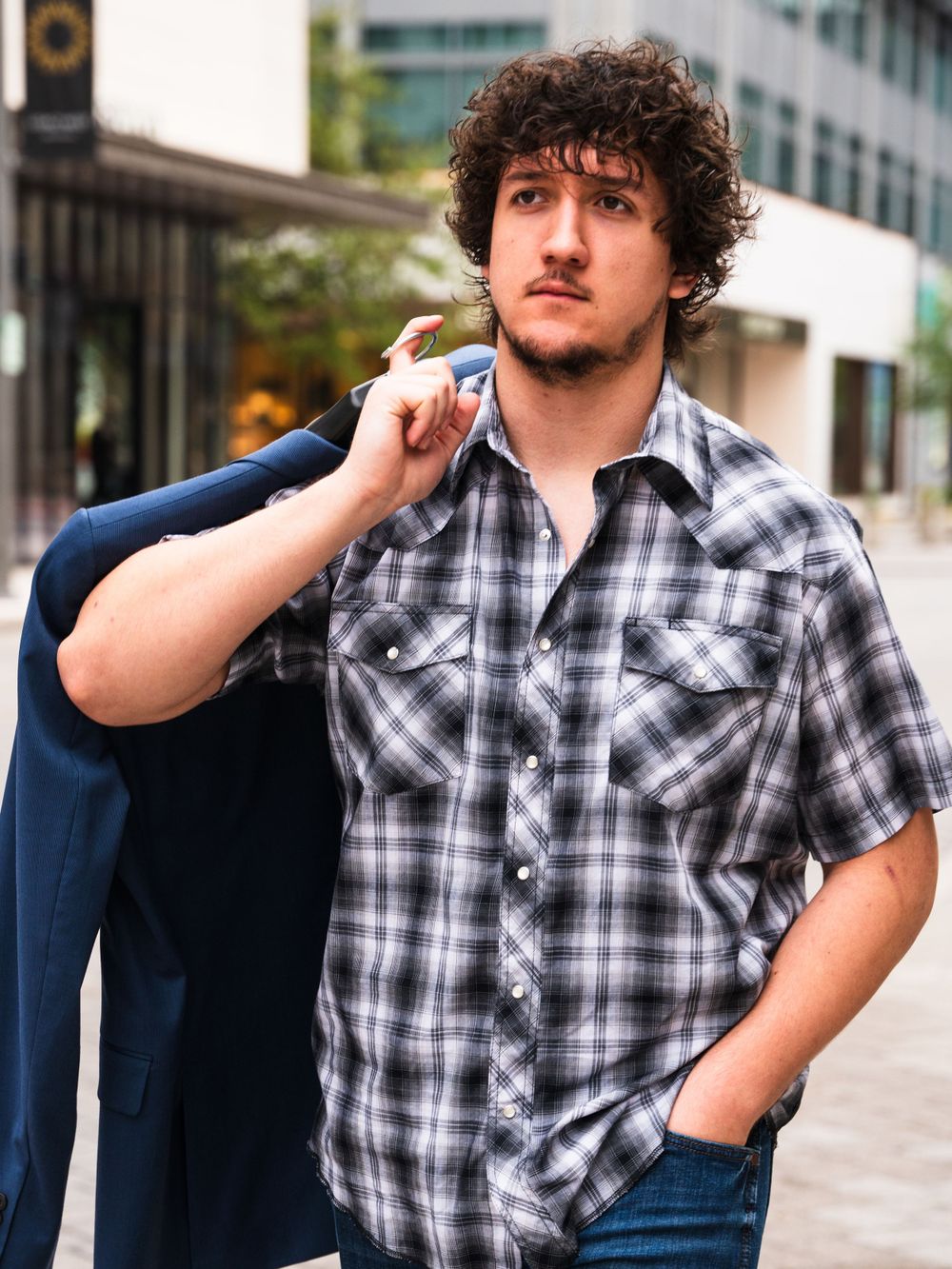 Person in plaid shirt carrying blue jacket over shoulder in urban setting.