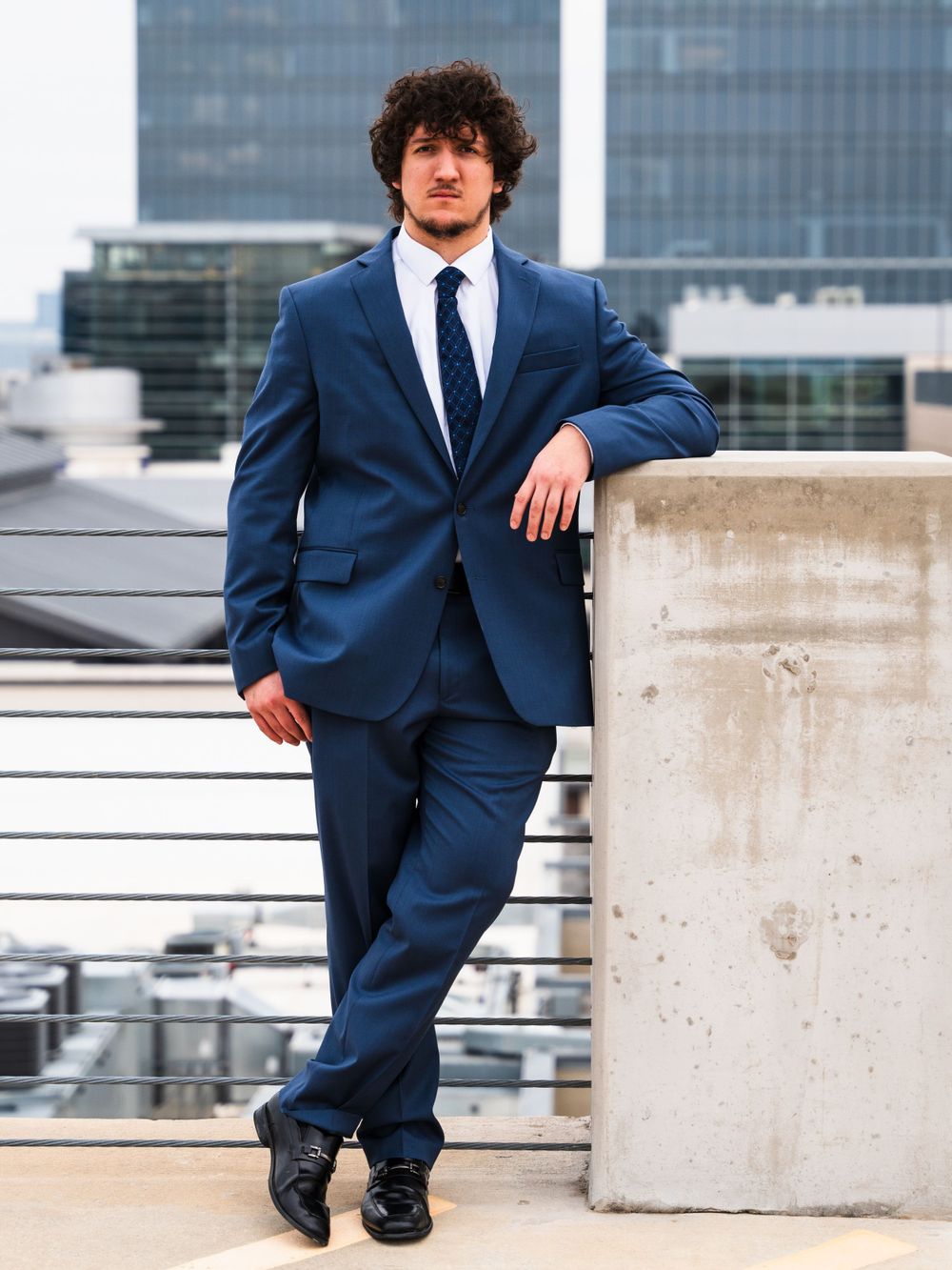 Professional portrait in navy suit leaning against concrete wall on rooftop.