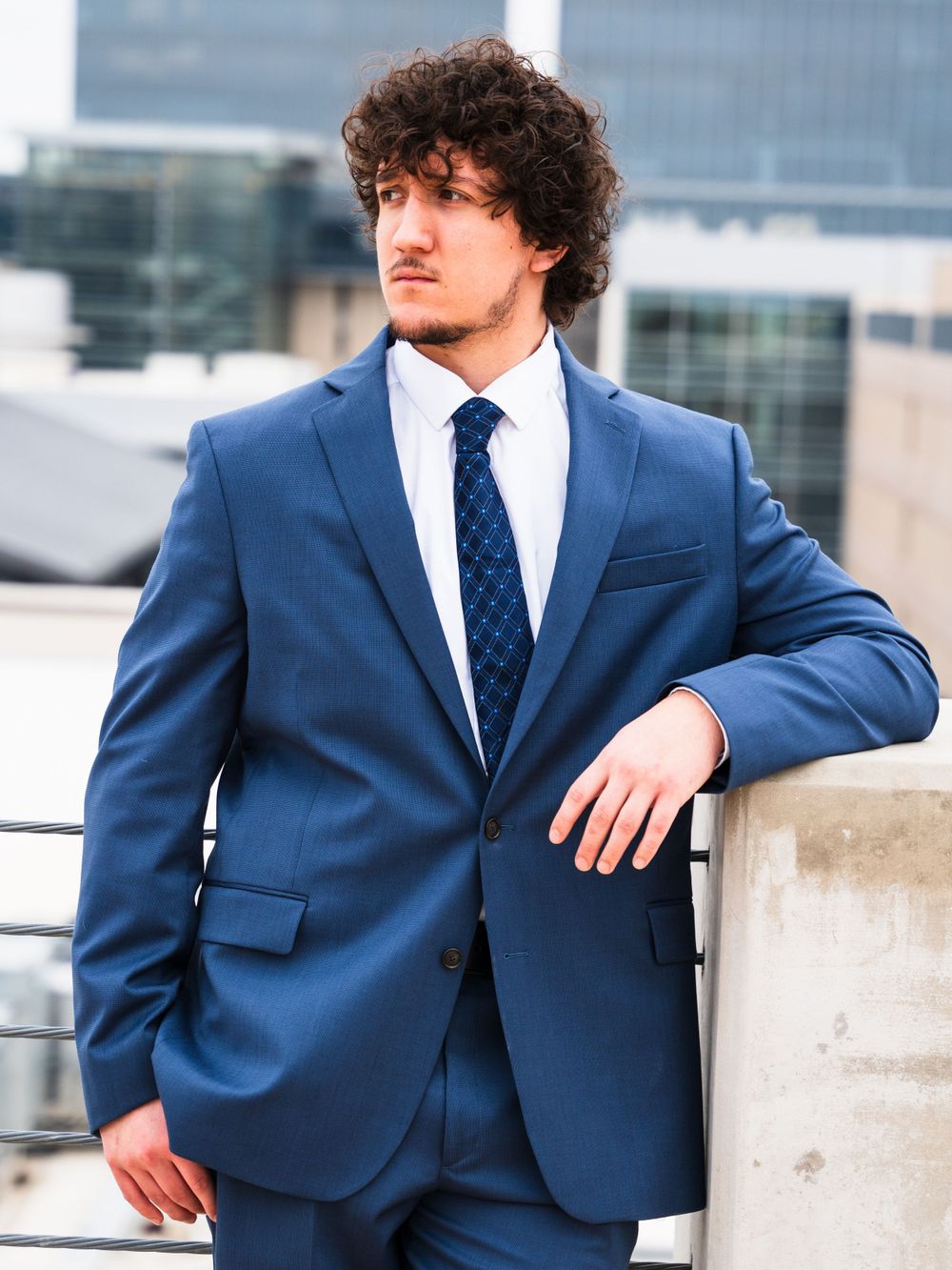 Formal portrait in fitted navy suit and polka dot tie against urban backdrop.