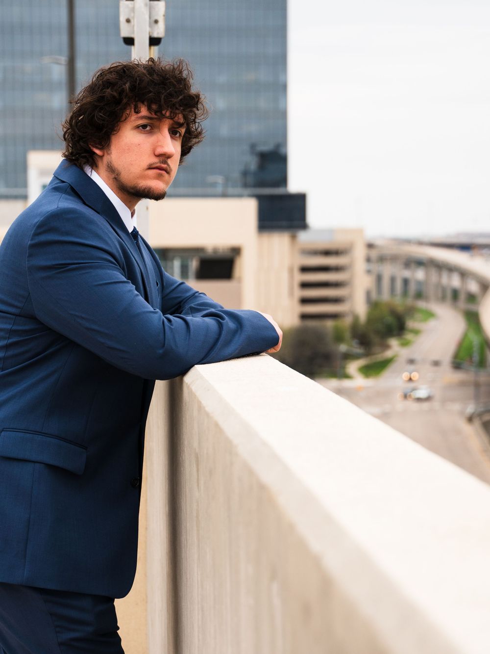 Professional in navy suit leaning on rooftop wall overlooking city landscape.