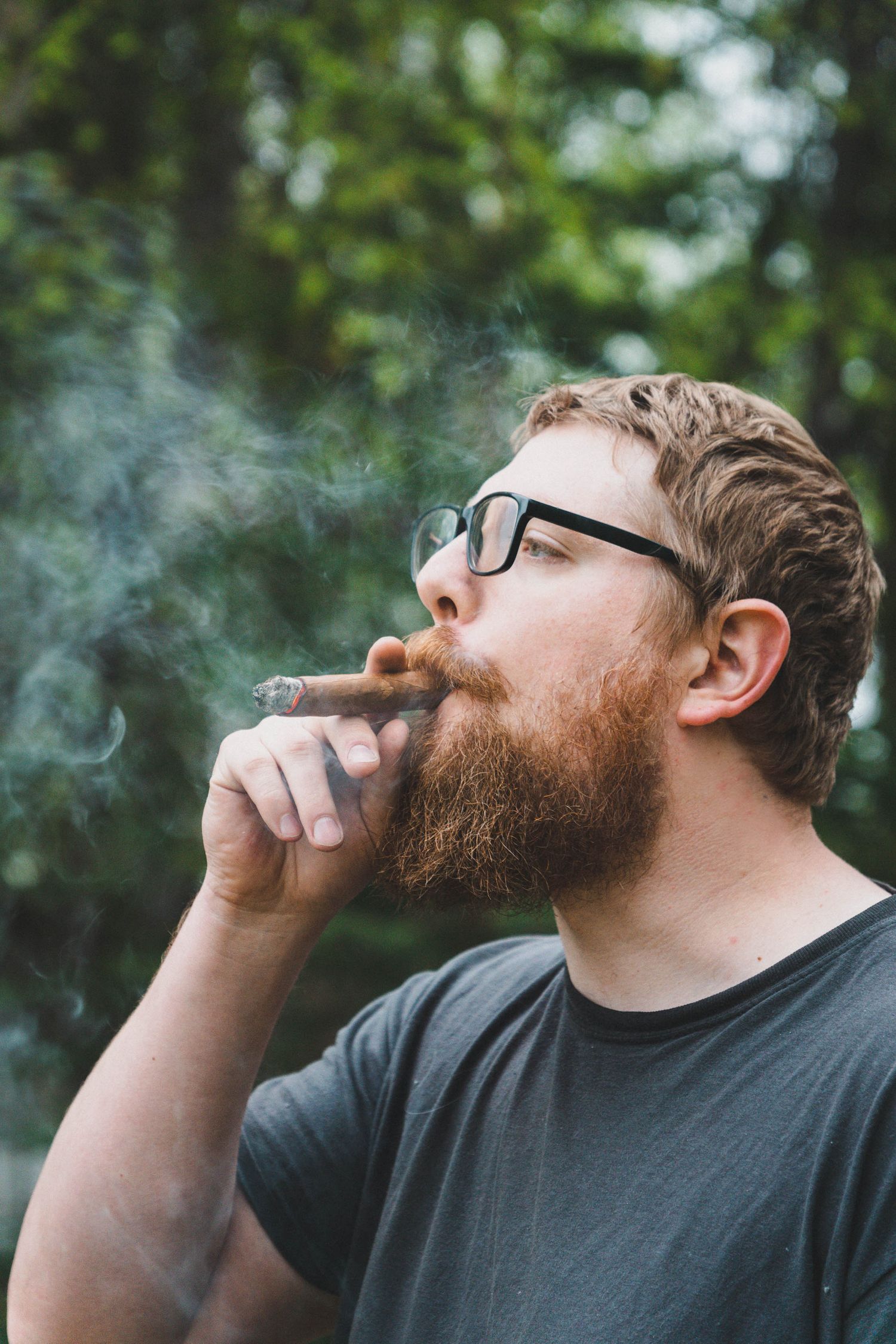 Bearded man with glasses smoking a cigar outdoors surrounded by green trees.