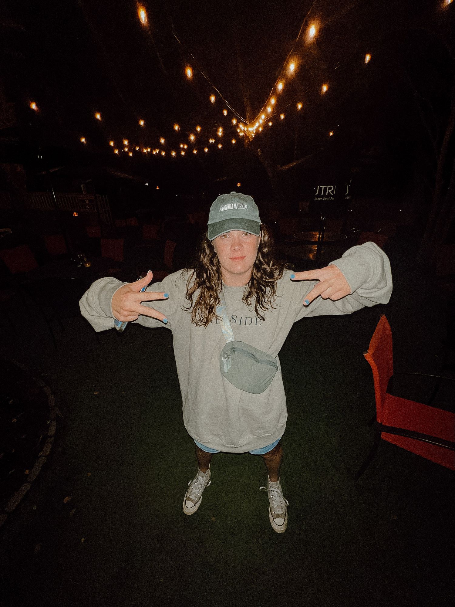 A figure poses under string lights at night wearing a sage green cap, white sweatshirt and sneakers.