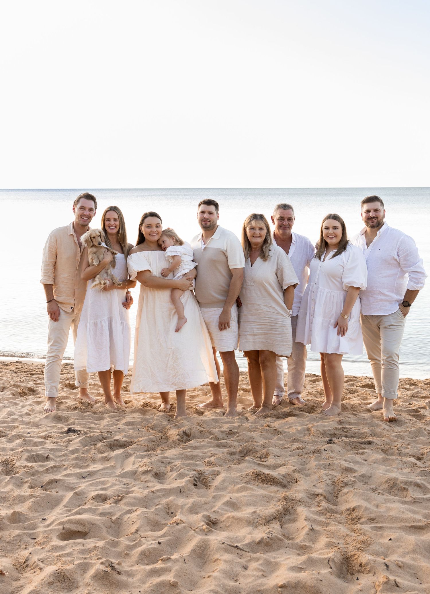 Group wearing coordinated white and beige outfits poses on a sandy beach at sunset.