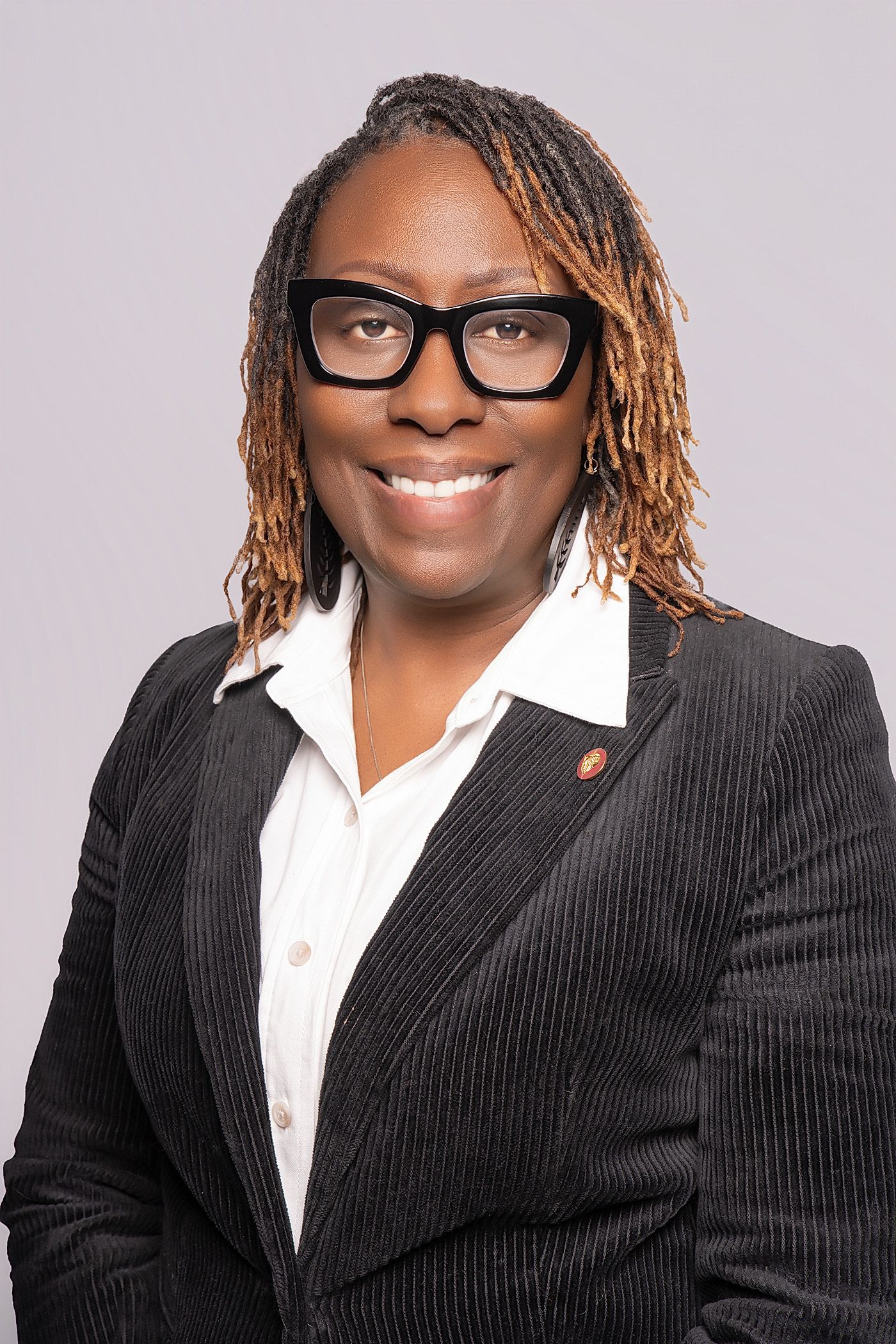 Smiling woman with golden-tipped locs, black-framed glasses, and a black pinstripe blazer poses in a professional headshot.