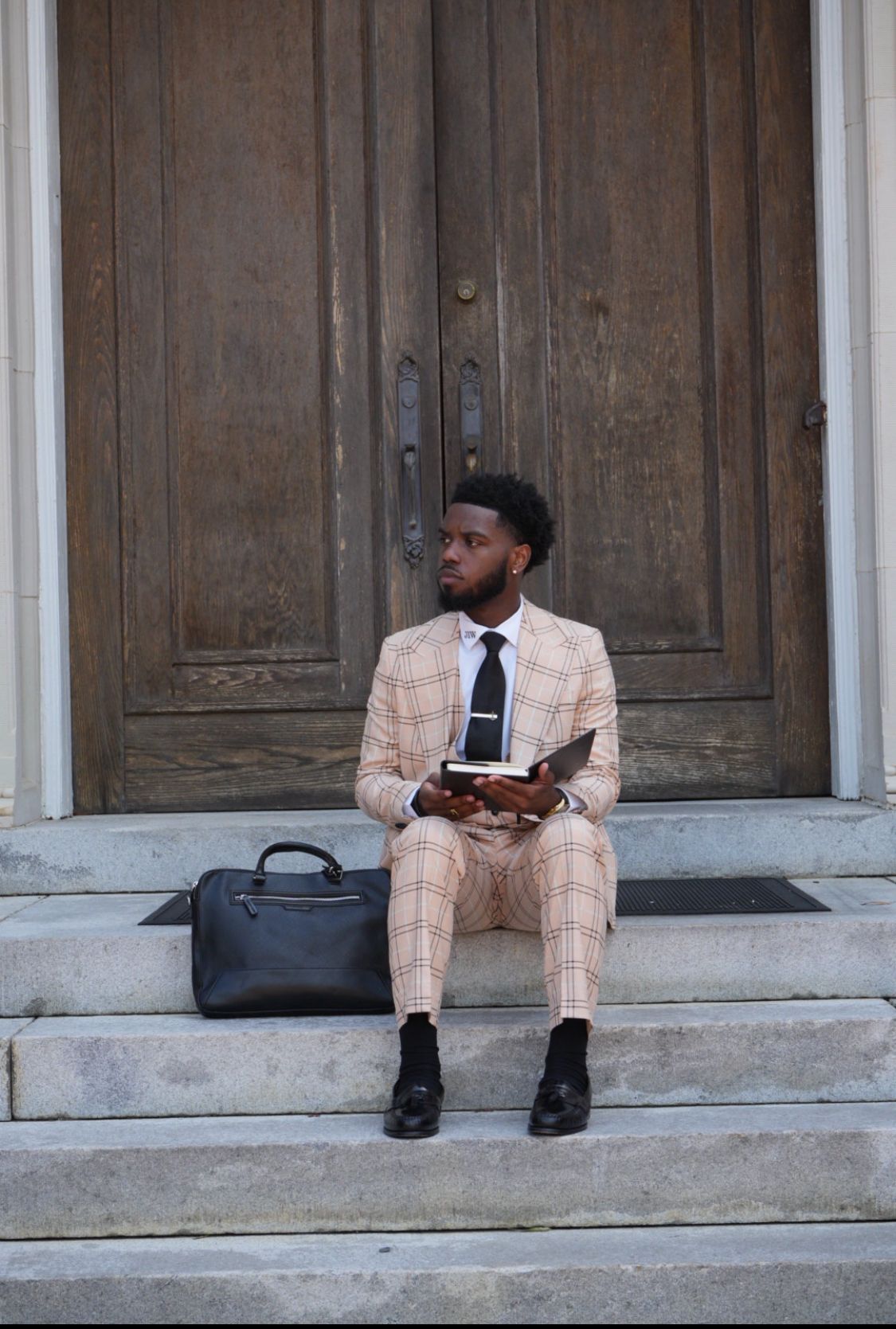 Man in a tan plaid suit and black tie sits on stone steps in front of a large wooden door, holding a book beside a black leather briefcase.