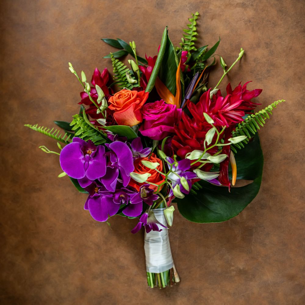 Tropical wedding bouquet with purple orchids, red roses, and green ferns wrapped in white ribbon.