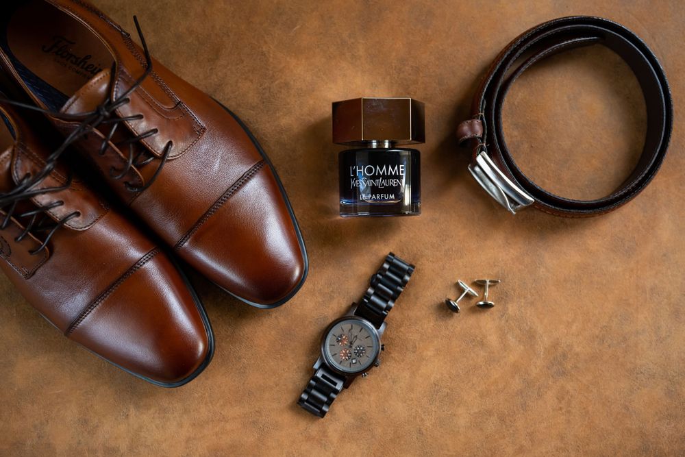 Groom's accessories including brown leather shoes, watch, cologne, and belt arranged on leather surface.