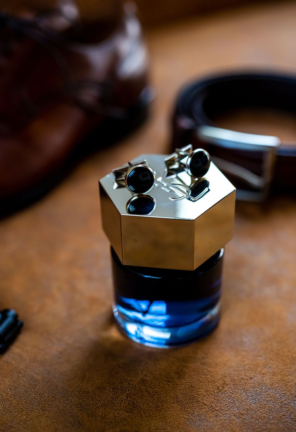 Blue cologne bottle with gold hexagonal cap displaying cufflinks on brown leather surface.