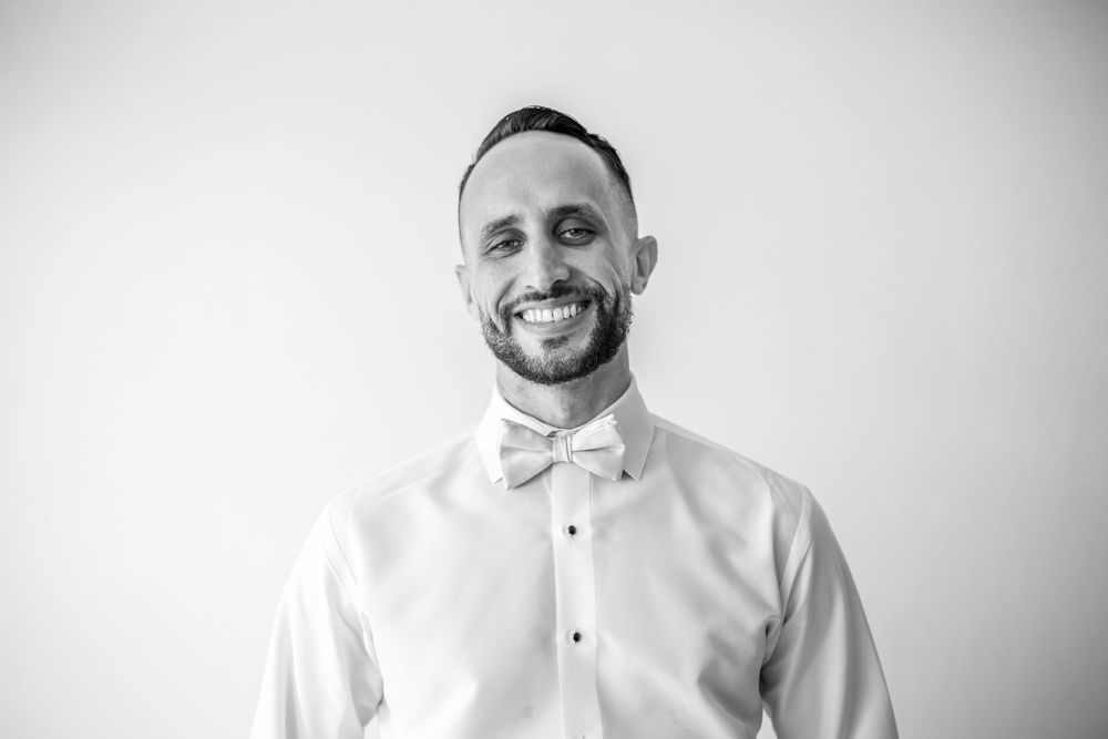 A smiling person wearing a white dress shirt and bowtie against light background.