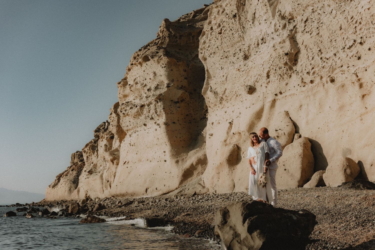 Bride and groom embracing on a beach with a tall cliff in Santorini