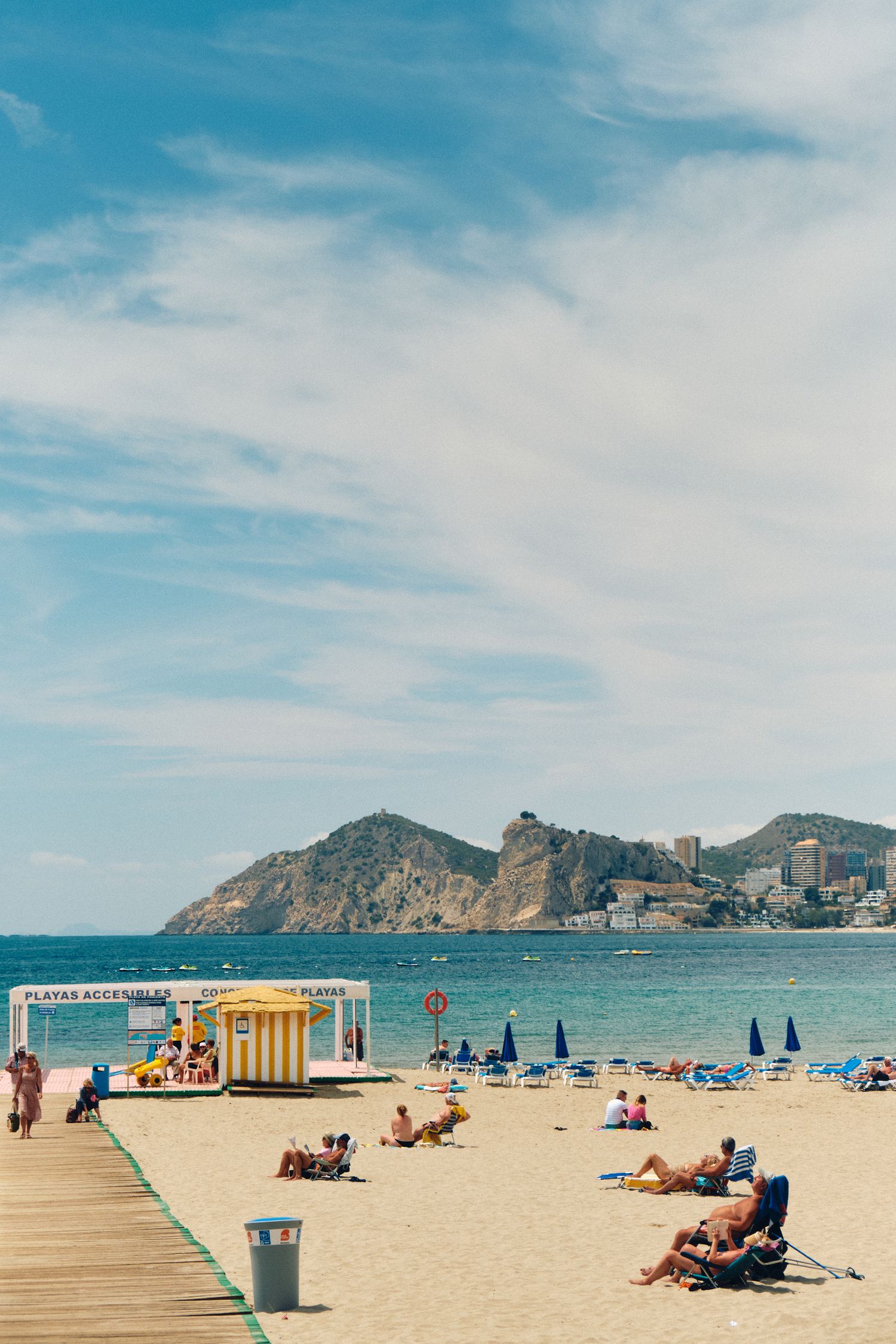 Wooden boardwalk and sandy beach with mountains and high-rise buildings in the background.
