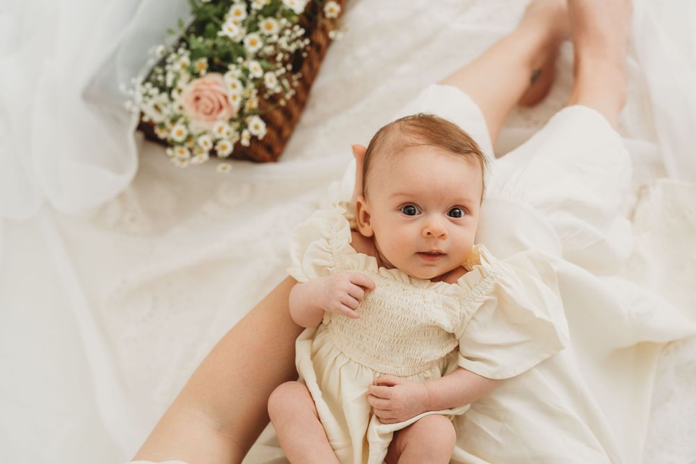 Baby in vintage white dress lying on soft fabric with flower bouquet in background.