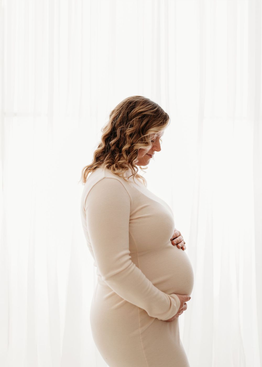 Side profile of pregnant woman in beige sweater against white curtains.