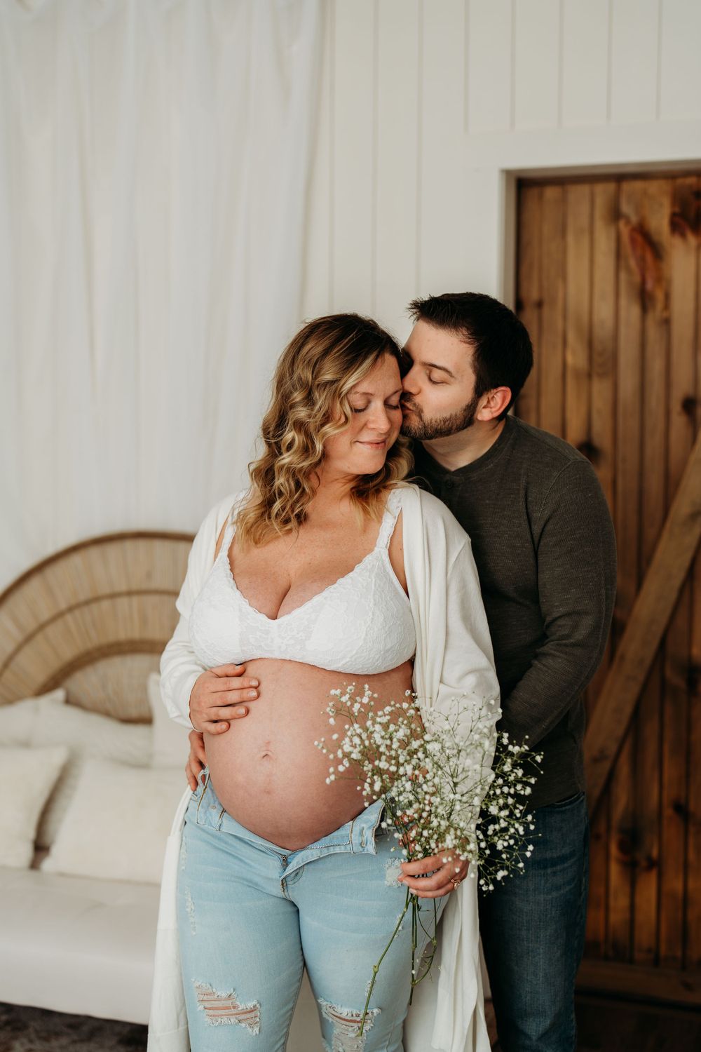 Intimate maternity photo featuring baby's breath flowers and ripped jeans in a minimalist indoor setting.