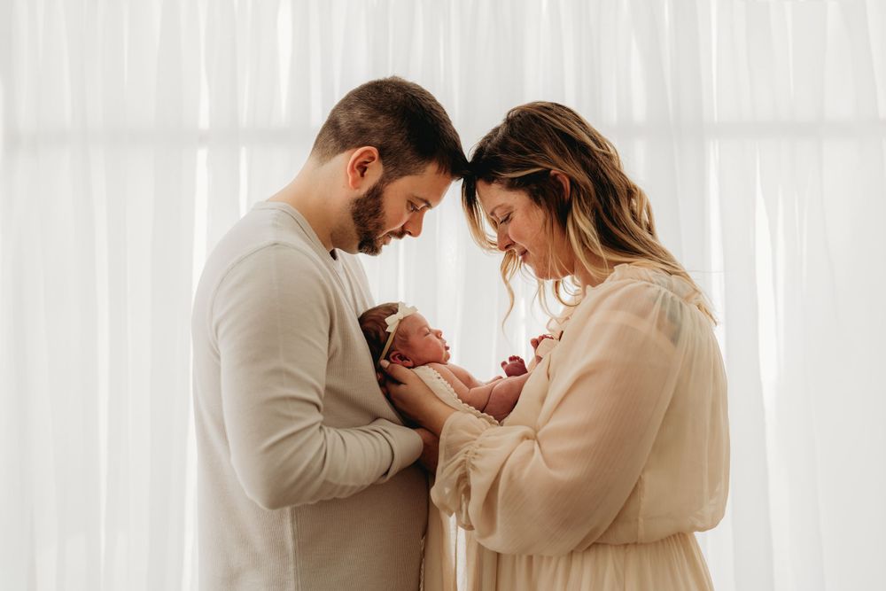Parents in neutral tones gazing lovingly at their newborn by window.