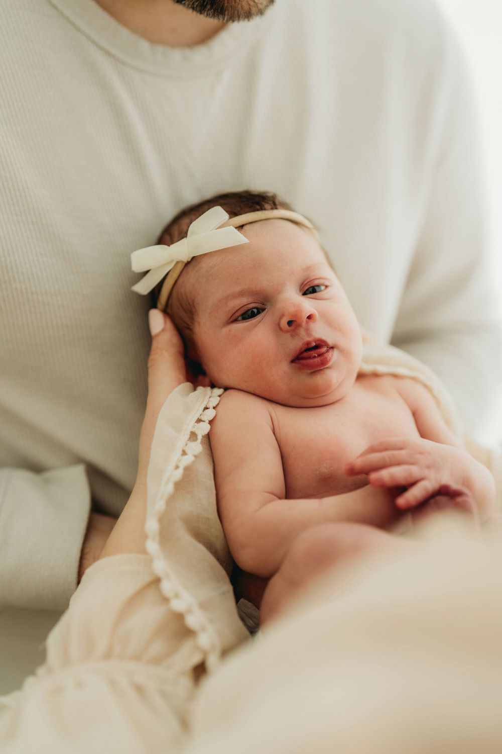 Sleeping newborn wearing white bow headband held against white sweater.