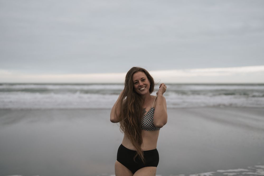 Woman in black swimwear poses joyfully on a cloudy beach with waves crashing in the background.