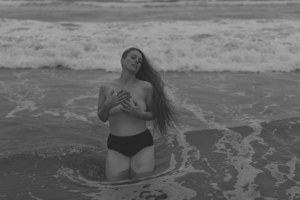 Figure kneeling in black bikini bottoms with long flowing hair poses in ocean waves at beach in black and white photo.