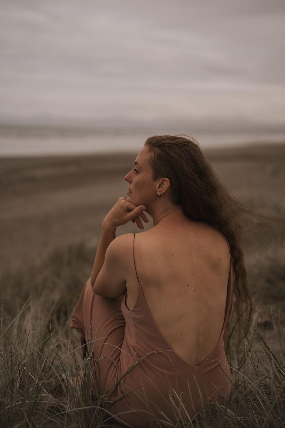 Woman in brown dress sits thoughtfully in tall grass under moody sky at sunset.