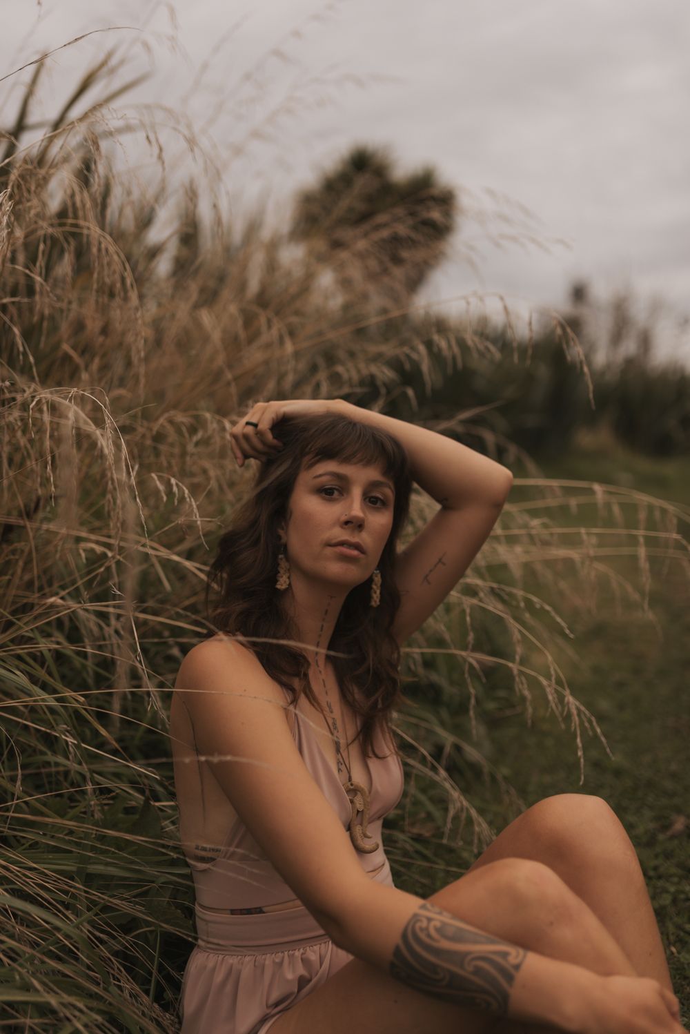 A woman in a beige dress poses thoughtfully amid tall prairie grass during golden hour lighting.