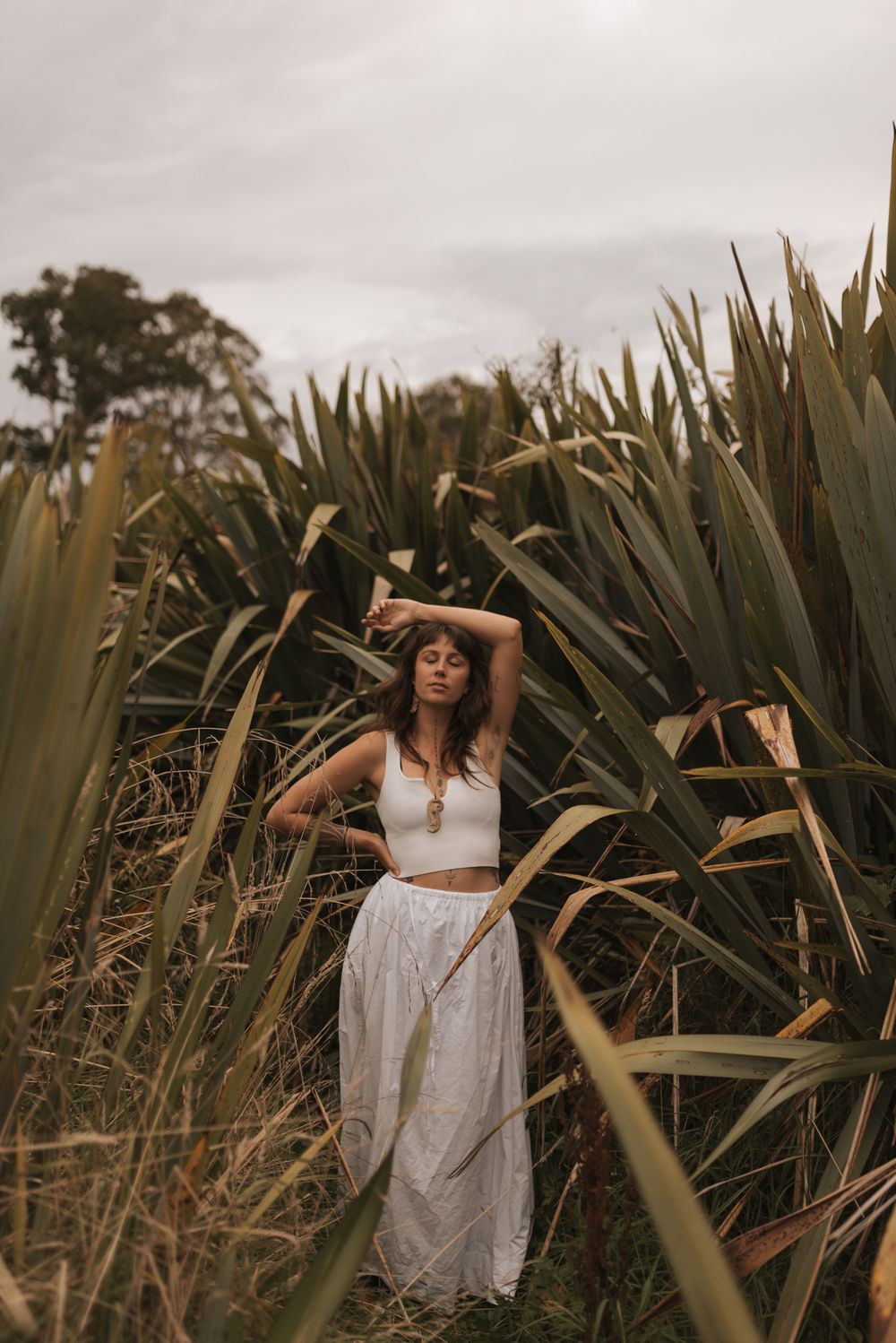 A person stands amid tropical foliage wearing a white crop top and flowing skirt in a natural outdoor setting.