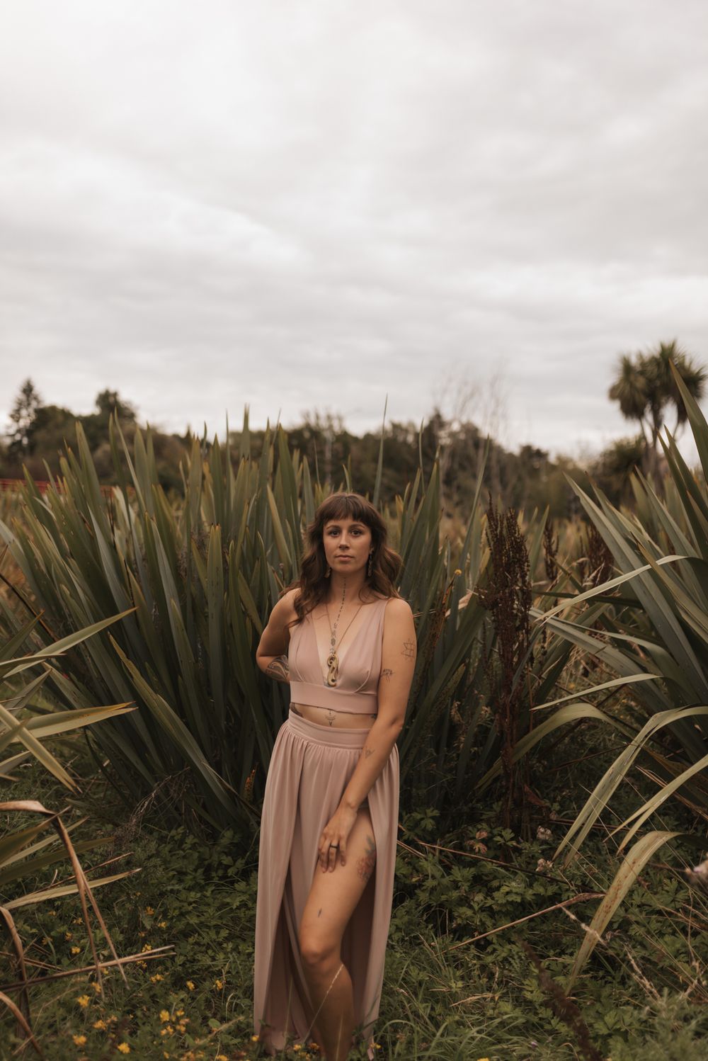 A model in a flowing beige dress poses among tall flax plants during a moody outdoor photoshoot.