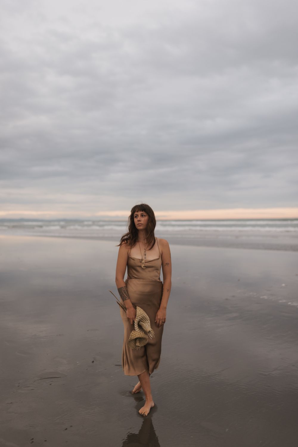 A person in a brown dress walks along a misty beach at dusk as waves roll in along the shore.