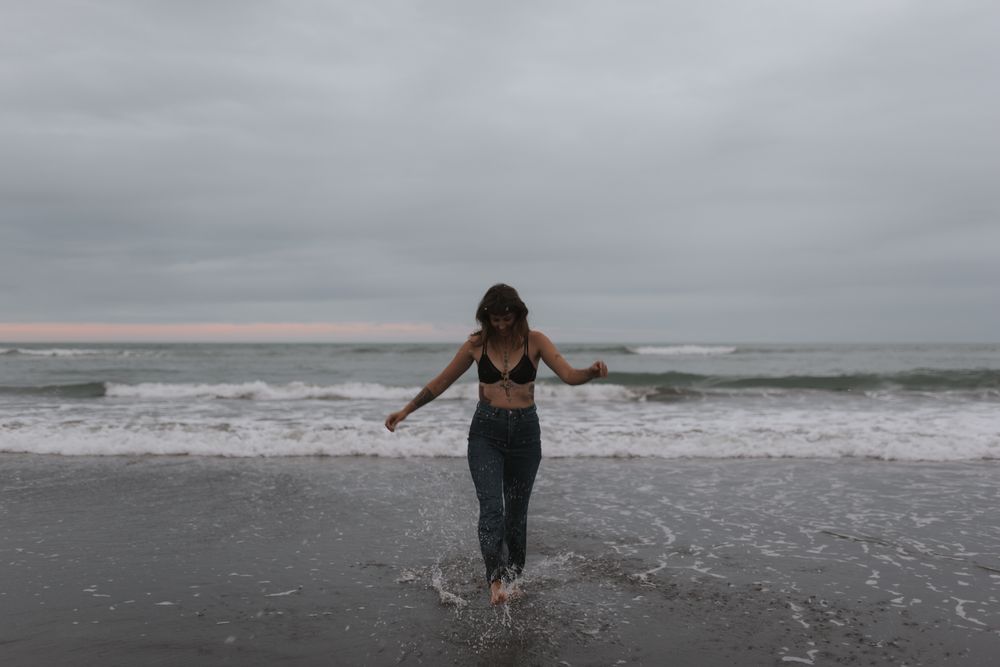 Silhouetted figure in a crop top and jeans splashes through waves on a moody beach under cloudy skies.