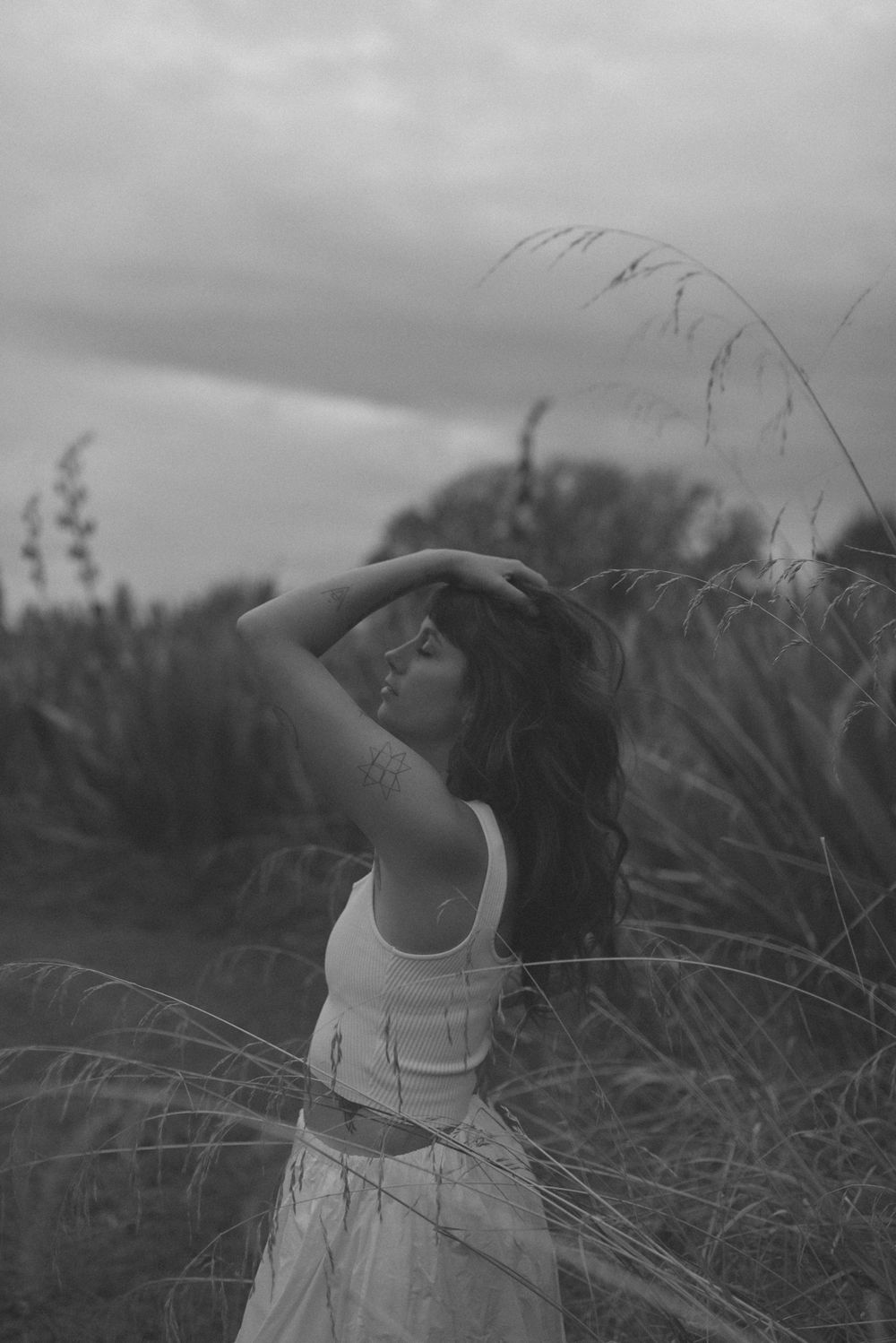 A figure in a white outfit poses dramatically in a field of tall grass under moody gray skies.