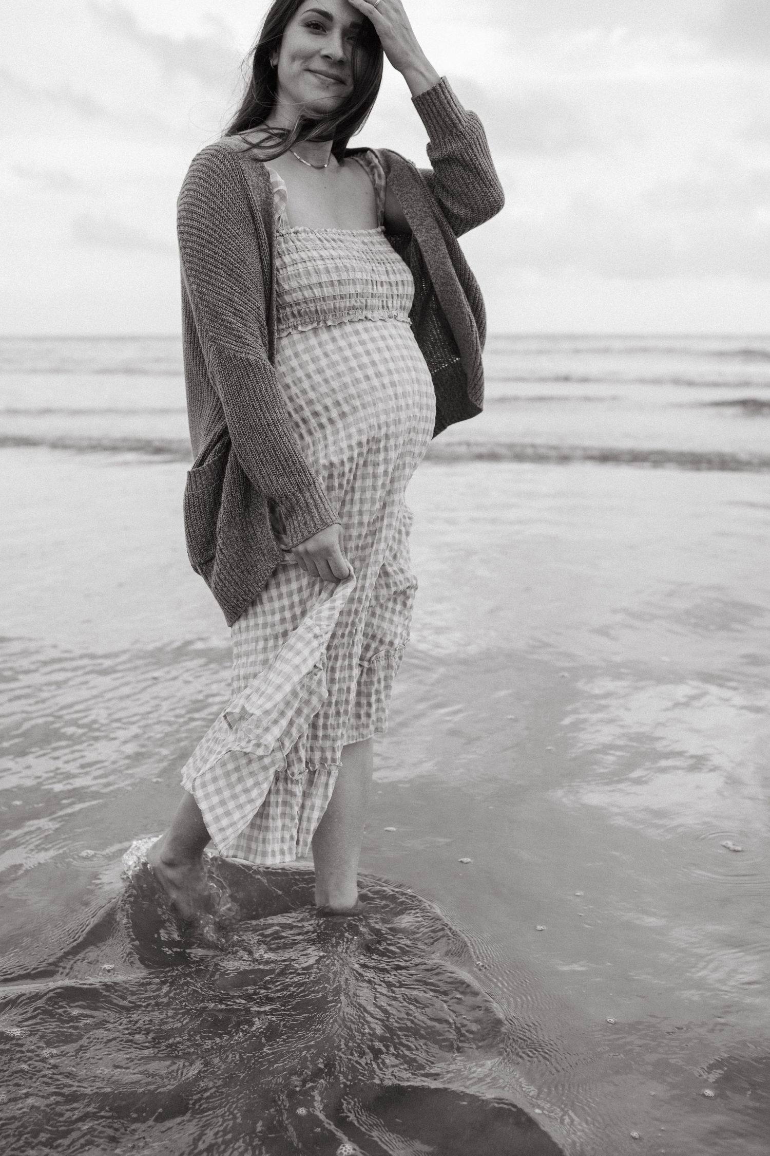 Expectant mother in gingham dress stands at ocean's edge in black and white photo during maternity photoshoot in San Diego.