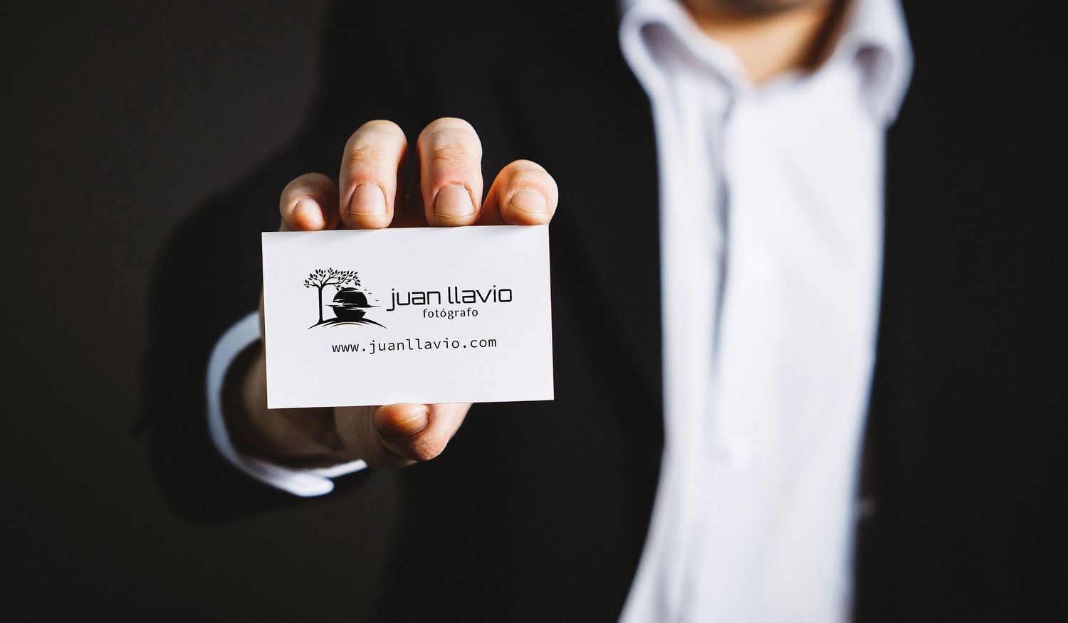 Mano en traje de negocios sostiene una tarjeta de presentación de Juan Llavio blanca sobre un fondo oscuro.