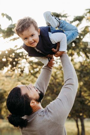 A father joyfully lifts his smiling toddler high in the air outdoors with sunlit trees in the background.