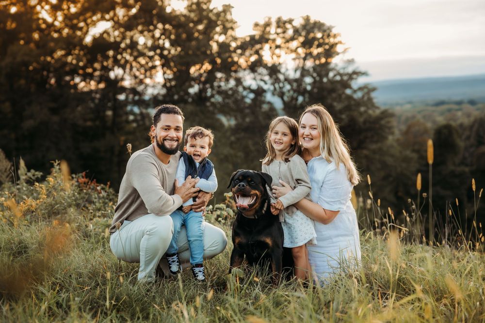 Family with two kids and black Labrador posing outdoors in golden hour sunlight among tall grass and trees.