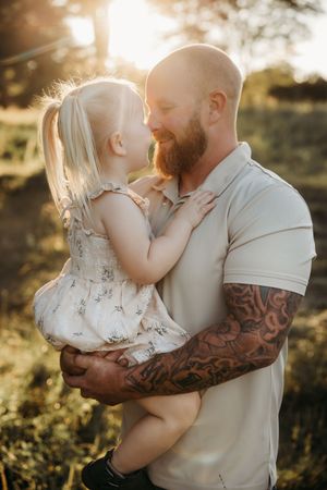 Tattooed father holding young blonde girl in floral dress, sharing tender moment in golden sunset light outdoors.