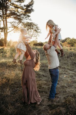 A joyful family lifting two young girls in a sunlit field during golden hour, wearing casual country-style outfits.