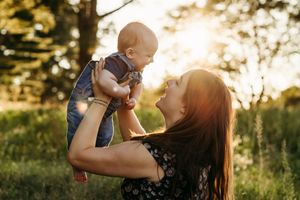 Mother lifting smiling baby in sunlit park, golden hour glow, joyful bonding moment outdoors.