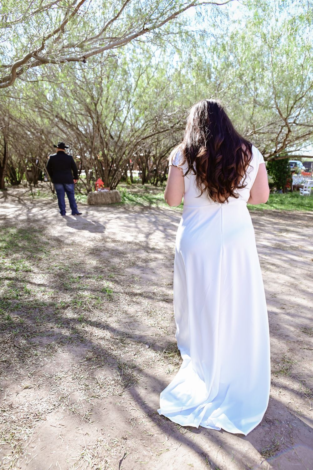 A white dress flows along a dirt path between trees as two figures walk in dappled sunlight.