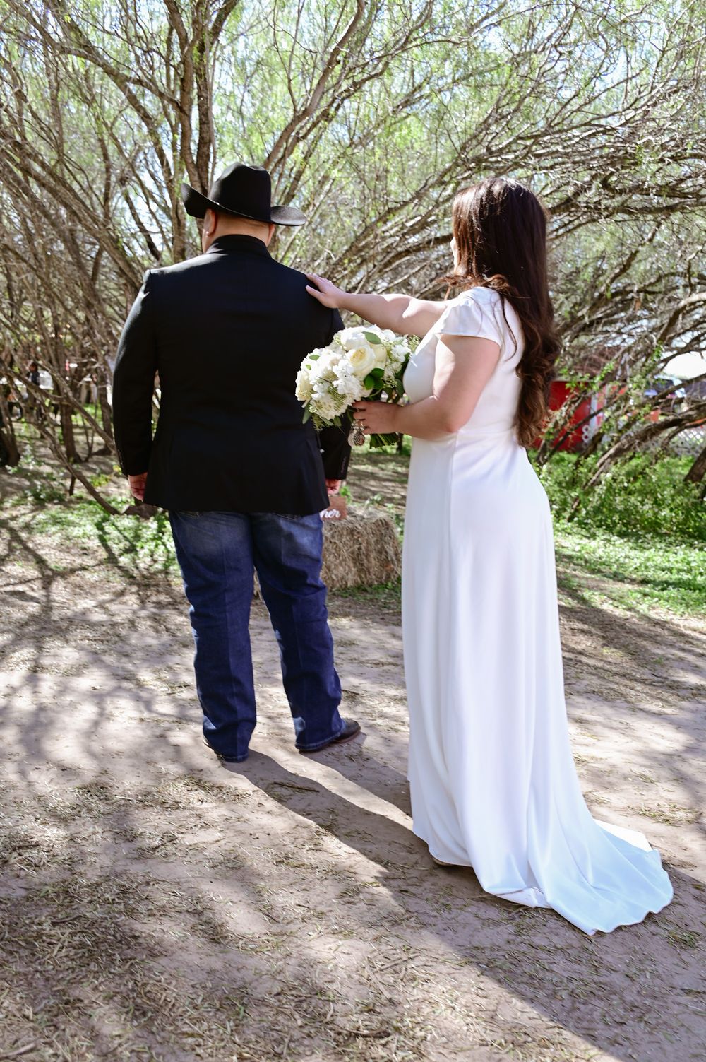 A couple in wedding attire walks along a tree-lined path holding a white floral bouquet.